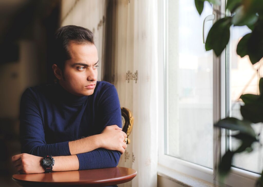 Man in a blue sweater leaning on a table and looking thoughtfully out a window, reflecting on his life patterns