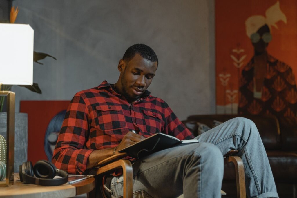 Man in a checkered shirt writing in a notebook, recording triggers and patterns for self-discovery