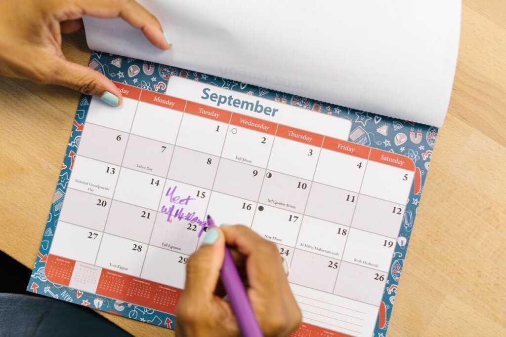 Hand marking a desk calendar with a purple pen, tracking daily progress toward a goal
